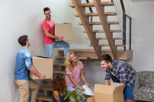 A group of five people, including three men and two women, are engaged in a house moving activity inside a residential home with a modern staircase made of wood and metal visible in the background. The individuals appear to be carrying and organizing cardboard boxes, some of which are open and contain household items. One man is standing on the staircase, holding a large cardboard box, while another man is on the ground, preparing to lift or move a box. The woman with blonde hair is sitting on the step below the staircase, smiling and possibly assisting with packing or organizing. The other woman, with curly red hair, is seated on the floor near the staircase, reaching towards the boxes. The scene takes place in a well-lit, contemporary interior with neutral wall colours and visible furniture, indicating an active house removals process with careful handling of household belongings, relevant to the services provided by Wandsworth Man with Van in the Wandsworth area. The environment suggests an informal yet organized residential move within a UK home.