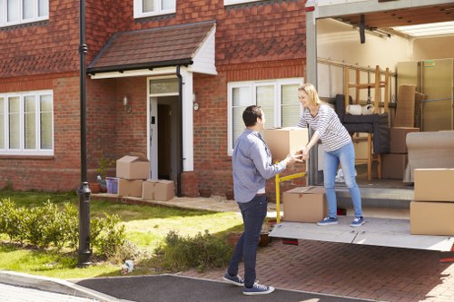 In the driveway of a residential property in Wandsworth, two individuals are engaged in a house removals task. A woman with blonde hair, dressed in a striped top and blue jeans, is standing inside a moving van with its back doors open, handing over a small cardboard box to a man with short dark hair, wearing a light blue shirt and dark jeans, who is standing outside on the driveway. The van is filled with various household items, including large cardboard boxes, a rolled-up rug, and pieces of furniture such as a black sofa, a wooden dining table, and chairs stacked inside. The house, constructed with red brick, features white-framed windows and a small porch over the front door. The surrounding area includes a tidy lawn with some small flower beds, a lamppost, and a paved street, suggesting a quiet residential neighbourhood in Wandsworth. The scene captures the moment of loading or unloading belongings, with natural daylight illuminating the scene clearly, emphasizing the textures of the boxes, furniture, and residential setting.