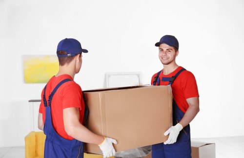 Two professional house removals workers, dressed in red t-shirts, blue overalls, and matching blue caps, are shown inside a modern, well-lit residential space. They are carefully lifting a large, rectangular cardboard box together, with one worker facing the camera and smiling, while the other has their back to the camera. The worker facing the camera is wearing white gloves. In the background, a white wall features a small abstract painting with yellow hues, and to the left, a fabric-covered yellow piece of furniture or cushion is partially visible. The setting appears to be a living room or hallway, with a clean, minimalistic interior, emphasizing the focus on the moving process. The scene captures a moment of careful handling and teamwork during a local Wandsworth house removal, highlighting the professional service provided by Wandsworth Man with Van. The environment is bright and tidy, indicating an organized domestic space typical of house relocation scenarios in the Wandsworth area.