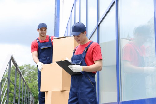 Two removal workers from Wandsworth Man with Van are outdoors, standing beside a modern building with reflective glass windows. The worker in the foreground, wearing a navy cap, red T-shirt, and blue overalls, is holding a clipboard and appears to be checking or recording items. The second worker, also in a navy cap, red T-shirt, and blue overalls, is standing behind him, holding several cardboard moving boxes of varying sizes, which are unsealed and stacked vertically. The background features a partly cloudy sky and a metal railing, suggesting they are on a balcony or external walkway of a residential or commercial property in Wandsworth, London. The workers are dressed in casual but professional attire suitable for house removals, and the scene emphasizes careful documentation and handling of household items during a relocation process. The reflective glass of the building adds a modern urban context to the scene, aligning with professional removals services in the Wandsworth area, focused on house and property moves.