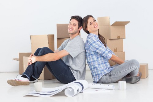 In a bright, minimalist room with a plain white wall background, a young man and woman are seated on the floor back-to-back amidst a collection of packed cardboard boxes of various sizes, some open and others sealed with packing tape. The man, wearing a light grey T-shirt and dark jeans, is smiling and has his arms crossed over his knees, while the woman, dressed in a blue and white checkered shirt with grey jeans, rests against him, looking slightly to the side with a relaxed expression. Around them, there are packing materials including a rolled-up sheet of brown paper, a white cup, and a small stack of papers. The scene suggests the process of moving house, typical of a professional removals service in Wandsworth or nearby South West London areas, with a focus on a smooth relocation experience. The well-lit environment emphasizes the organized packing stage of a household move, with no furniture or household appliances visible, highlighting the preparatory phase of a house removals service by Wandsworth Man with Van.