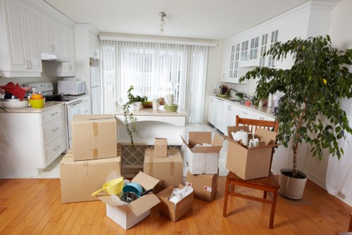 A well-lit kitchen in a Wandsworth home with white cabinetry and a wooden floor, featuring several open cardboard boxes filled with various household items such as plastic containers, kitchen utensils, and paper goods. Some boxes are stacked on each other, while others are on the floor or on a wooden chair. In the background, large windows with vertical blinds allow natural light to fill the space, revealing a white dining table with a potted plant and a bowl on top. To the right side, a tall potted plant with leafy branches extends near the wall, adding greenery to the room. The scene captures a moment of packing or relocation preparation, typical of house removals activities in South West London, with removal equipment and packed items ready for moving, reflecting the process managed by Wandsworth Man with Van.