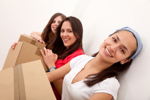 Three women in a well-lit interior space, possibly a house hallway or room, are smiling and appear to be preparing for a move. The woman in the foreground wears a white top and a blue headscarf, holding a cardboard box with her right hand while facing the camera. Behind her, two other women, one in a red top and one in a dark top, are also holding boxes and smiling. The boxes are made of brown corrugated cardboard and vary in size, with some visible packing tape on them. The background is plain white, suggesting a clean, organized environment typical of a domestic setting in Wandsworth or nearby areas. The scene reflects the process of house removals or moving services, with the women’s expressions conveying positivity and teamwork. The arrangement of the boxes and the presence of the women suggest they are either packing or carrying items in preparation for a move, typical of services offered by Wandsworth Man with Van within the local London borough of Wandsworth.