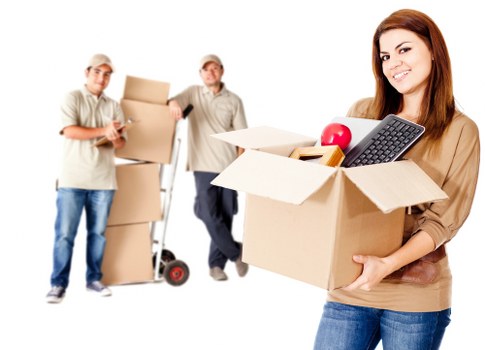 A young woman with long brown hair, wearing a beige top and blue jeans, is smiling while holding an open cardboard moving box filled with items such as a red apple, a calculator, and a notebook, in a well-lit indoor setting. Behind her, three men are engaged in packing and moving household belongings: one man is holding a smartphone and has a small stack of cardboard boxes; another man, dressed in a light-colored shirt, glasses, and jeans, is standing next to a hand truck loaded with stacked boxes; the third man, also dressed casually, is leaning on the hand truck, partially obscured. The scene depicts a professional house removal process, with the background suggesting a home environment and the focus on packing and transportation activities, consistent with services offered by Wandsworth Man with Van in the Wandsworth area within London.