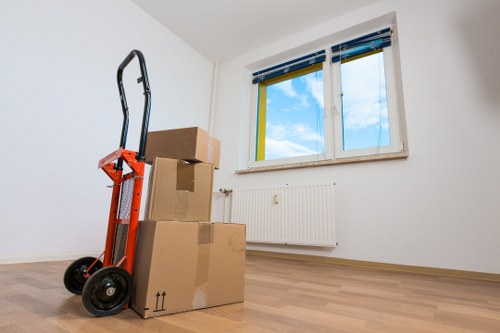 An empty room with white walls and light wooden flooring, featuring a white radiator beneath a double-glazed window with blue and yellow window frames showing a partly cloudy sky outside; in the foreground, a red hand truck with a black handle and wheels leans against a small stack of three cardboard boxes of varying sizes, all sealed with brown packing tape, indicating preparation for moving within the Wandsworth area; the room appears to be part of a residential property in London, typical of house removals services offered by Wandsworth Man with Van, with natural daylight illuminating the space and minimal furnishings present.