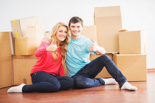 A young couple sitting on a wooden floor in a well-lit room, surrounded by stacked beige cardboard moving boxes of various sizes, some sealed with packing tape, with the woman wearing a coral pink long-sleeved top and dark jeans, and the man in a light blue T-shirt and dark jeans; both are smiling, giving a thumbs-up gesture, indicating a successful house move. The background shows a plain white wall, suggesting an indoor residential setting, possibly in Wandsworth or nearby in South West London, consistent with professional removals services by Wandsworth Man with Van. The scene captures a moment of satisfaction after packing or loading, with the floor’s warm wood tone contrasting with the light tones of the boxes and the casual clothing of the individuals.