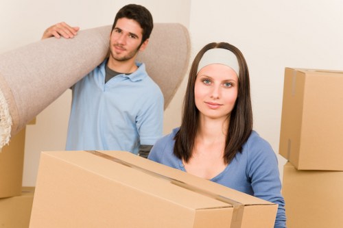 A young man and woman, both with fair skin and casual attire, are depicted in a moving or packing scenario inside a residential setting. The man, wearing a light blue shirt and with short dark hair, is standing in the background, lifting a large, light grey upholstered armchair by its backrest, preparing to move or position it. The woman, with straight brown hair and a neutral expression, sits in the foreground, holding a medium-sized cardboard box with visible folded flaps, likely filled with household items for a house removal. Several other cardboard boxes of various sizes are placed around them, some stacked and others positioned on the floor, indicating an active packing process. The setting appears to be a well-lit room with neutral walls, and the background suggests a typical residential interior, possibly in Wandsworth or nearby in South West London. The scene captures the practical aspects of a house removal, with attention to their clothing, the textures of boxes, and furniture, complemented by the natural lighting typical of a daytime move, reflecting the services offered by Wandsworth Man with Van in the area.