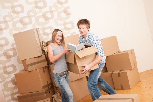A young man and woman are standing in a room with a light-colored wall featuring a geometric pattern, surrounded by a number of cardboard moving boxes of various sizes. The woman, on the left, has shoulder-length blonde hair and is wearing a sleeveless grey top with ripped jeans; she is smiling and holding an open book or magazine. The man, on the right, has short, curly brown hair and is dressed in a checked shirt with rolled-up sleeves and blue jeans; he is lifting a medium-sized cardboard box with both hands and appears to be smiling. The boxes are stacked against the wall and arranged on a wooden floor, some boxes are on the floor while others are placed on top of each other, indicating an ongoing or recent move within a residential property. The scene is well-lit, with natural or artificial light highlighting the details of the boxes, furniture, and their expressions, reflecting the typical activity of house removals in the Wandsworth area, with no visible furniture or appliances besides the boxes, all placed for transport or packing.