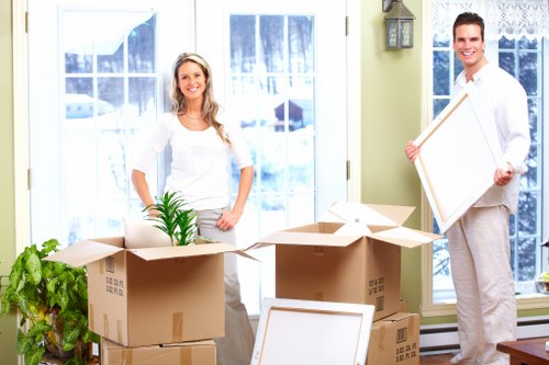 A smiling woman with long, wavy blonde hair wearing a white top and light grey trousers stands inside a bright, well-lit living room with large windows and lace curtains. She is surrounded by several open cardboard boxes, some filled with household items, including a small potted green plant in one of the boxes. To her left, a green houseplant is positioned beside a stack of sealed boxes. On the right, a man with short dark hair, dressed in a white shirt and light grey trousers, is holding a picture frame with a white border, preparing for a house move. The room features a neutral-toned wall with a small exterior lamp mounted beside the window, and the overall setting suggests a house in Wandsworth undergoing packing or unpacking in the process of a home relocation, consistent with local removals services provided by Wandsworth Man with Van.