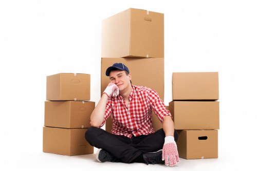 A young man with light skin, wearing a red and white checked shirt, dark trousers, a navy cap, and pink work gloves, is sitting cross-legged on the floor of a bright space with a plain white background. He is resting his head on his left hand and smiling gently. Surrounding him are multiple cardboard moving boxes of various sizes, some stacked with one large box positioned behind him, while smaller boxes are arranged to his left and right. The setting suggests an indoor area, possibly part of a house or flat, in the process of a move in Wandsworth. The boxes appear to be empty, prepared for packing or transportation, and the scene conveys a professional yet relaxed atmosphere typical of a house removals service. The arrangement of the boxes and the tidy environment subtly indicate a professional move, emphasizing the topics of house removals and relocation in the Wandsworth area.