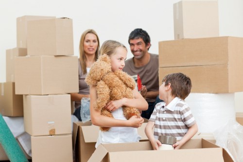 A family inside a bright room in a house, surrounded by stacked cardboard moving boxes in various sizes, some open and filled with household items. The mother and father are standing in the background, smiling and observing their children. In the foreground, a young girl with blonde hair, wearing a beige fuzzy cardigan and light blue jeans, is holding a brown teddy bear and talking to a boy wearing a striped polo shirt. The boy is sitting inside an open cardboard box, which also contains a small metallic object. The room features a clean, modern interior with white walls and natural lighting, possibly during a house move in the Wandsworth area. The scene captures the temporary clutter of a typical house removal process, emphasizing the preparation for a relocation service provided by Wandsworth Man with Van, with a focus on safety and organized packing in a residential setting.