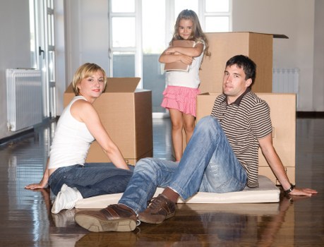 A family of three, comprising a woman, a man, and a young girl, seated on a wooden floor inside a residential property with light-colored walls and large windows in the background. The woman, wearing a white sleeveless top and blue jeans, is sitting on the floor with her legs extended forward and slightly bent, facing the camera. The man, dressed in a striped short-sleeve shirt and blue jeans, is leaning back on his hands with his legs stretched out, positioned to the right of the woman. The young girl, standing behind them, has shoulder-length blonde hair and is wearing a pink skirt with a white top; she is standing with her arms crossed and smiling. Surrounding them are several cardboard moving boxes, some open and partially filled, placed on the floor, suggesting a house removal or relocation process. The setting indicates an active moving environment with natural lighting from the windows emphasizing the household interior details, such as the wooden flooring and simple decor. The scene is natural, neutral, and professional, reflecting a typical family moving day within the Wandsworth area.