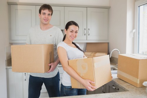 In a well-lit kitchen, a young man and woman are preparing to move house. The man, dressed in a light grey sweater and dark jeans, is holding a medium-sized cardboard box, while the woman, wearing a white t-shirt and blue jeans, is carrying a slightly larger box. Behind them, light grey kitchen cabinets with silver handles are mounted on the wall, with a beige cardboard box on the kitchen counter near a white sink. The window behind the woman provides natural daylight, illuminating the scene and highlighting the modern tiling and countertop. Several other cardboard boxes are placed on the kitchen countertop and floor, some sealed with packing tape, indicating an ongoing packing process typical of house removals within Wandsworth. The scene reflects a typical home environment preparing for a local move, with household items and moving supplies visible, aligning with professional removals services in the Wandsworth area, UK.
