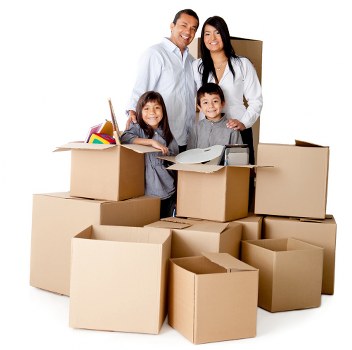 A family of four standing inside a room surrounded by numerous open and closed cardboard moving boxes, some filled with household items such as plush toys, a small radio, and plastic containers. The family members, consisting of two adults and two children, are smiling at the camera and standing close together, with the mother and father behind the children. The children are in front, one girl and one boy, holding a cardboard box filled with household items. The background shows a plain wall with natural indoor lighting illuminating the scene, suggesting a residential interior preparing for a move or unpacking. All boxes are made of brown Kraft material, varying in size and shape, with some open revealing contents. The scene reflects a typical house relocation scenario in Wandsworth, with clear focus on packing and household organization, integral to professional removals services by Wandsworth Man with Van.